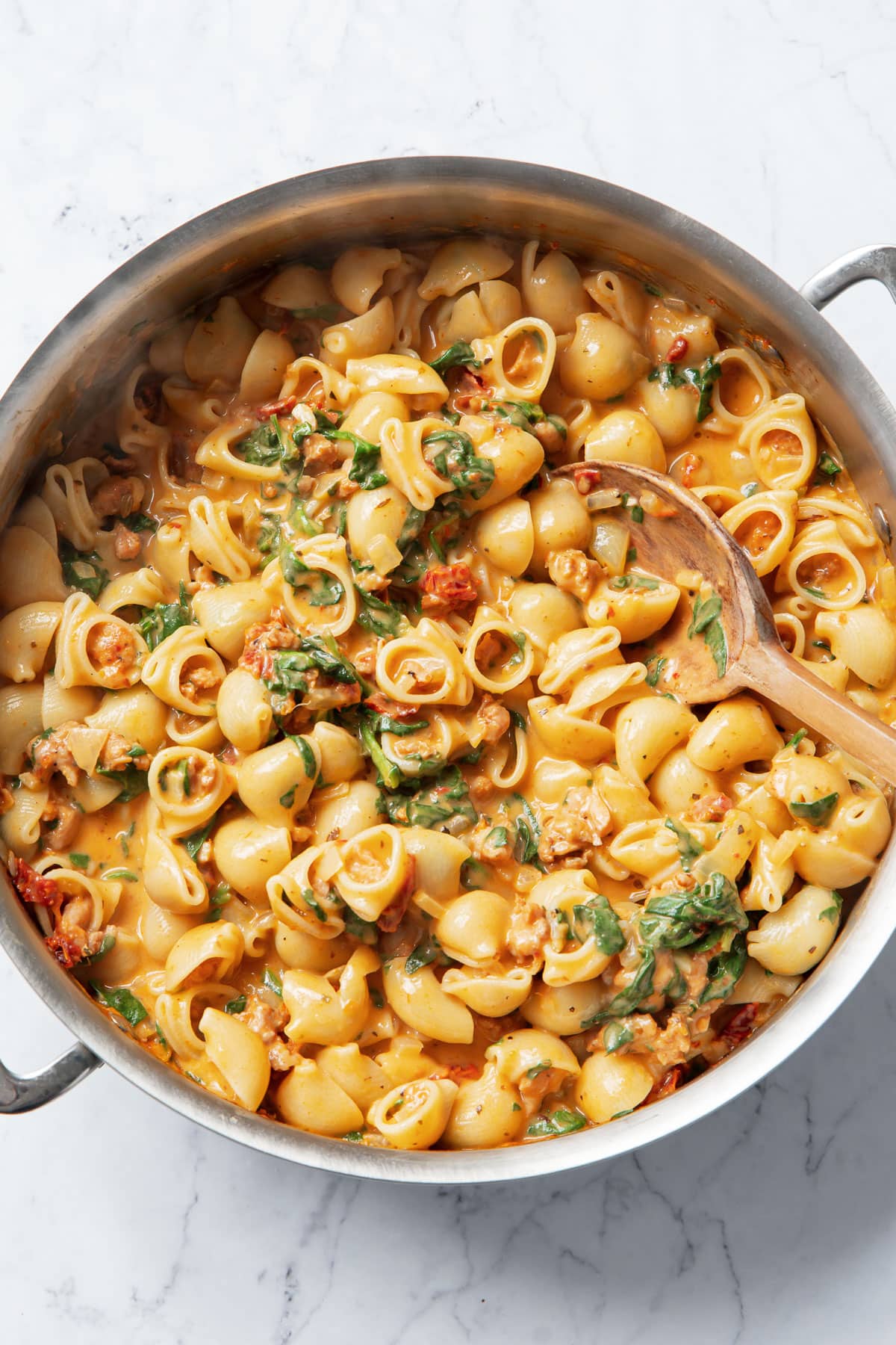 Overhead stainless steel saucepan filled with Creamy Sundried Tomato and Italian Sausage Pasta and a wooden spoon.