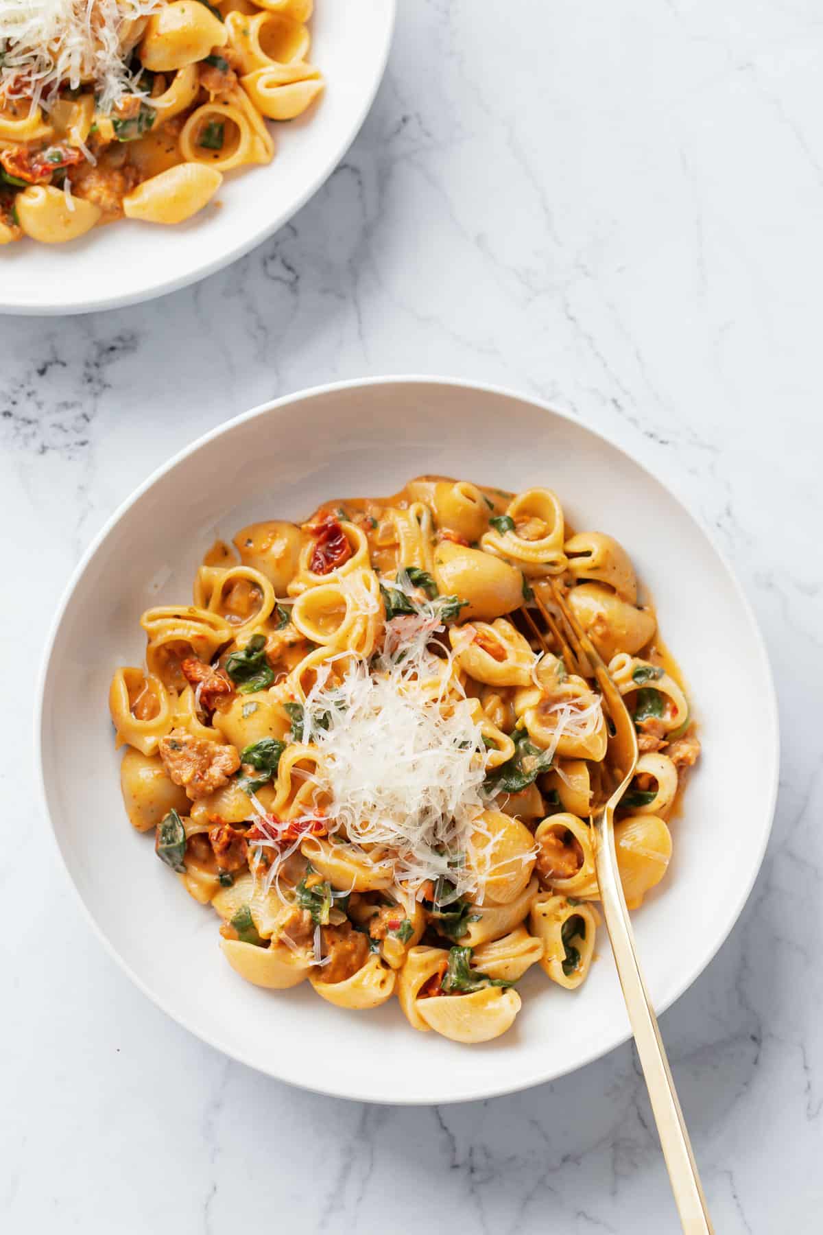 Overhead of two bowls of Creamy Sundried Tomato and Italian Sausage Pasta and topped with finely shaved parmesan cheese with gold forks on a marble background.