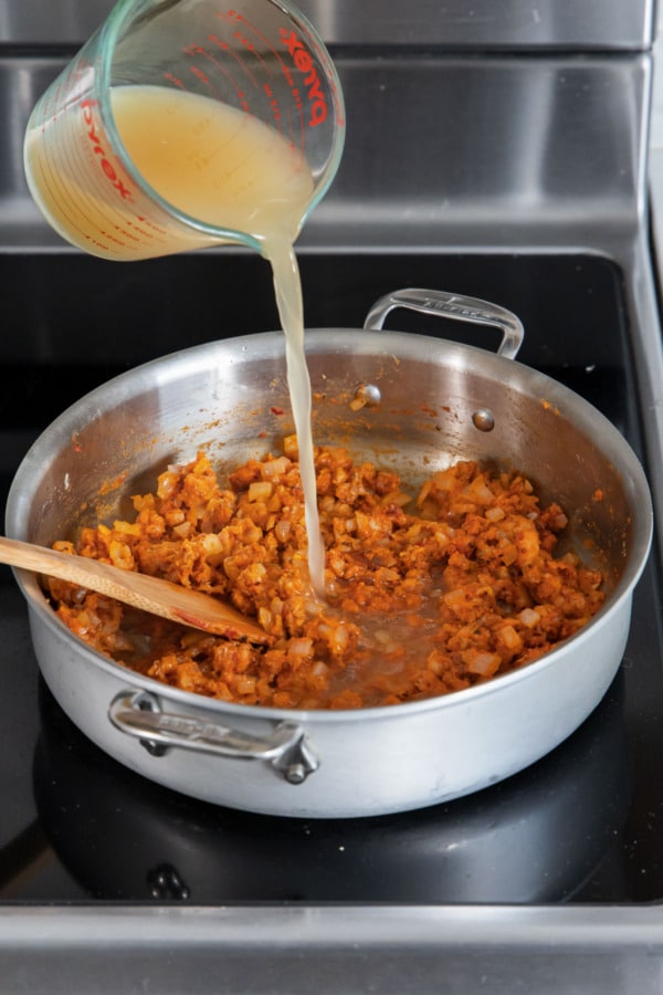 Adding 1.5 cups of chicken broth to a stainless skillet along with other ingredients.