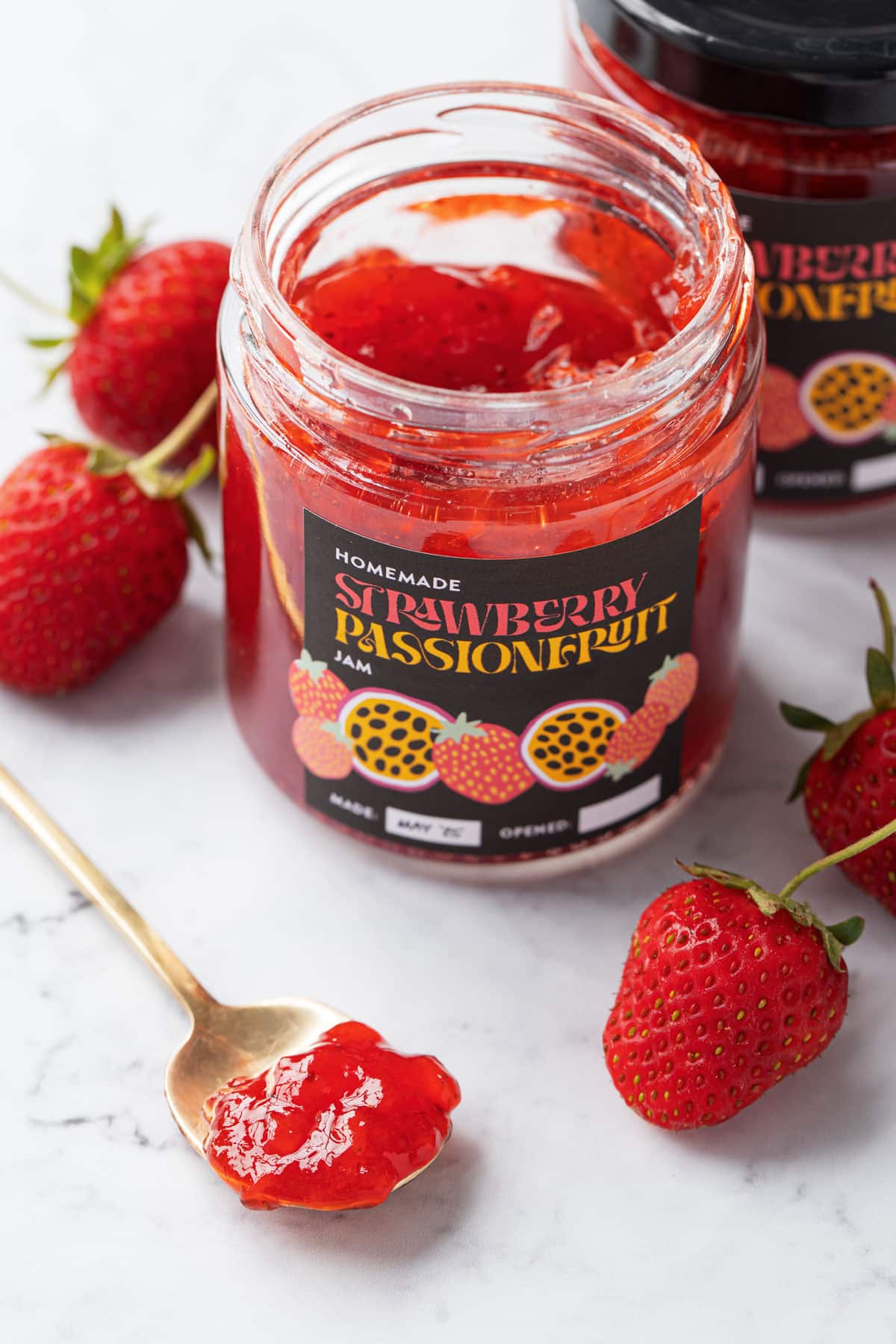Jar of Strawberry Passionfruit Jam on a marble background with a golden spoon filled with jam in the foreground to show the consistency of the jam.