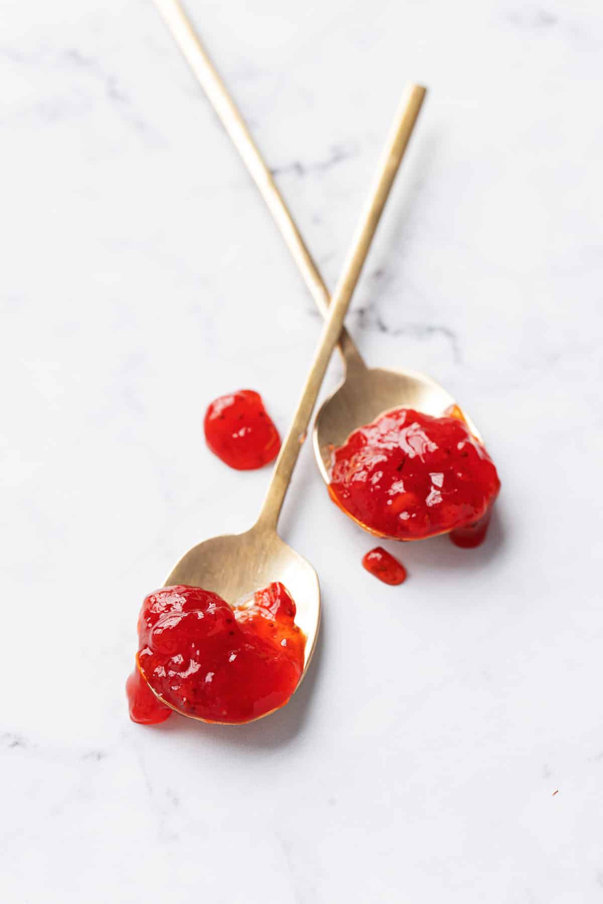 Two gold spoons, crossed on a marble background and filled with ruby red Strawberry Passionfruit Jam, a few messy dollops dripped onto the background.