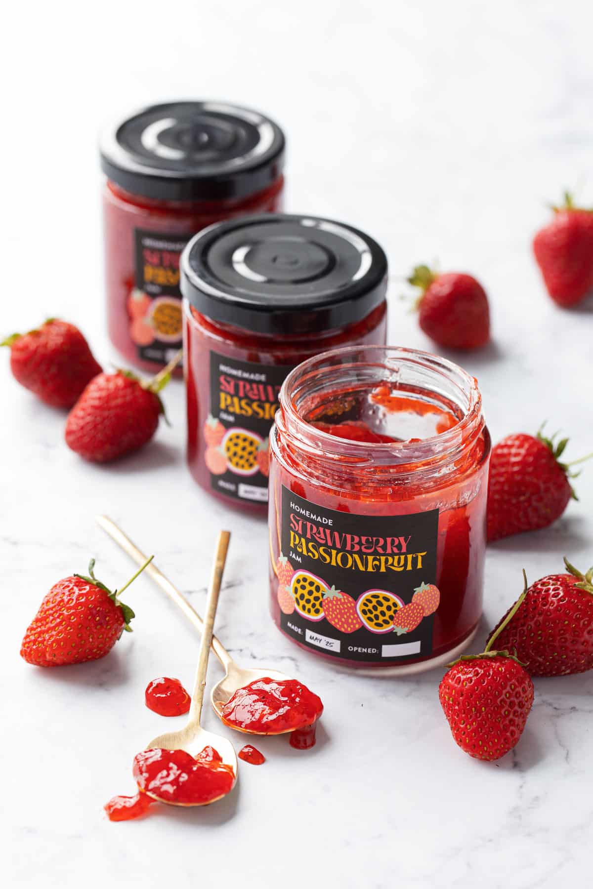 Three jars of Strawberry Passionfruit Jam on a marble background, with one jar open and a few spoonfuls of jam on the side.