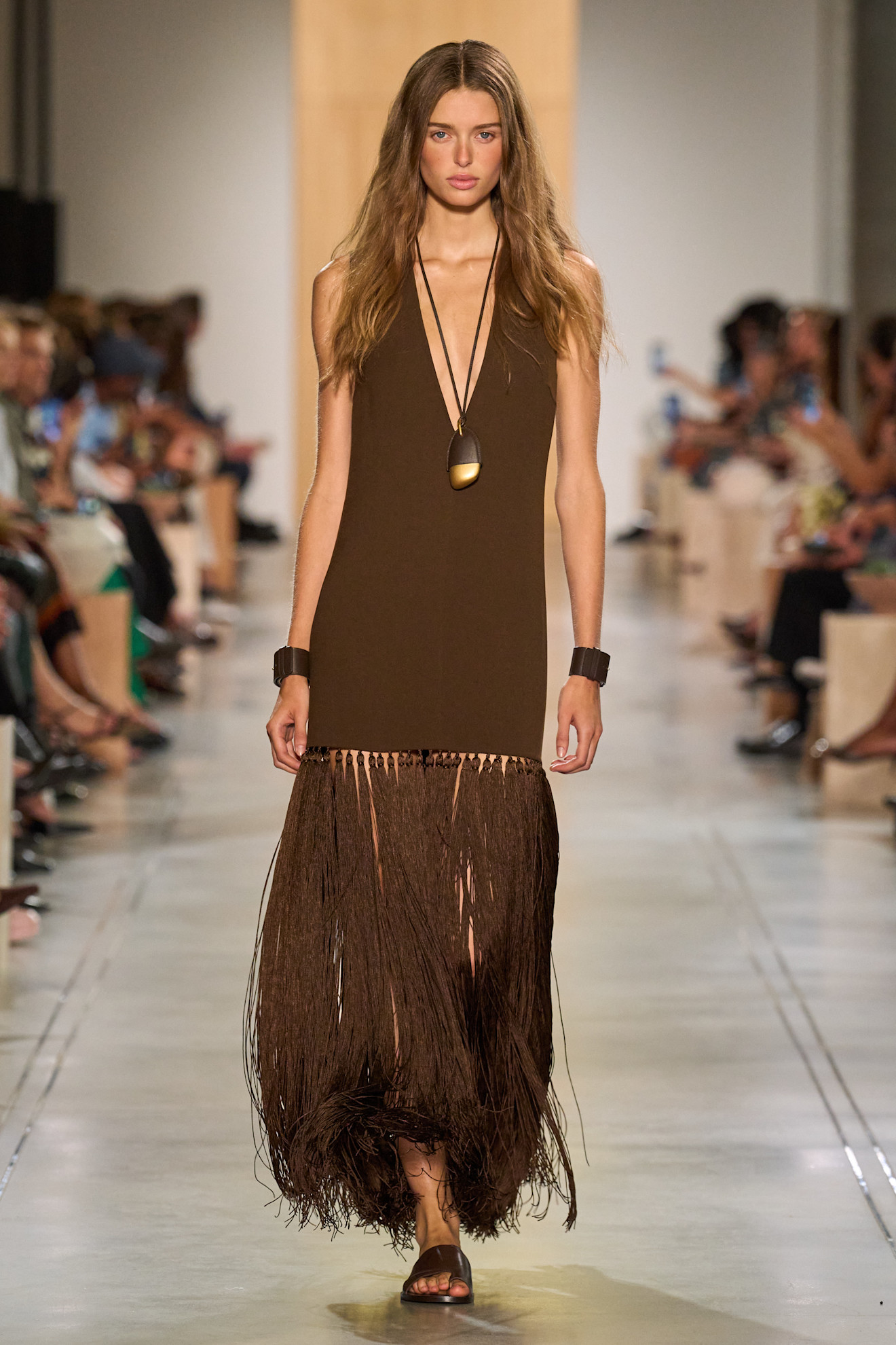 A Michael Kors model wearing a brown V-neck dress with a fringe hemline