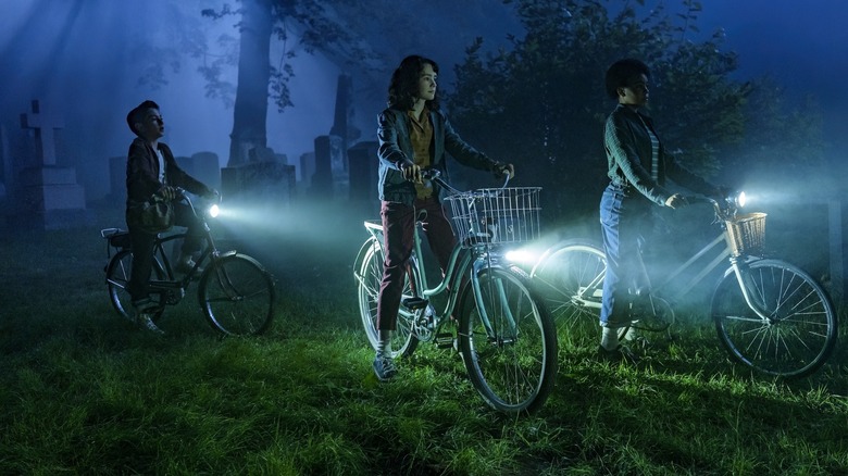 Arian S. Cartaya as Rich, Clara Stack as Lilly Bainbridge, and Amanda Christine as Ronnie Grogan riding bikes in a cemetery at night in It: Welcome to Derry