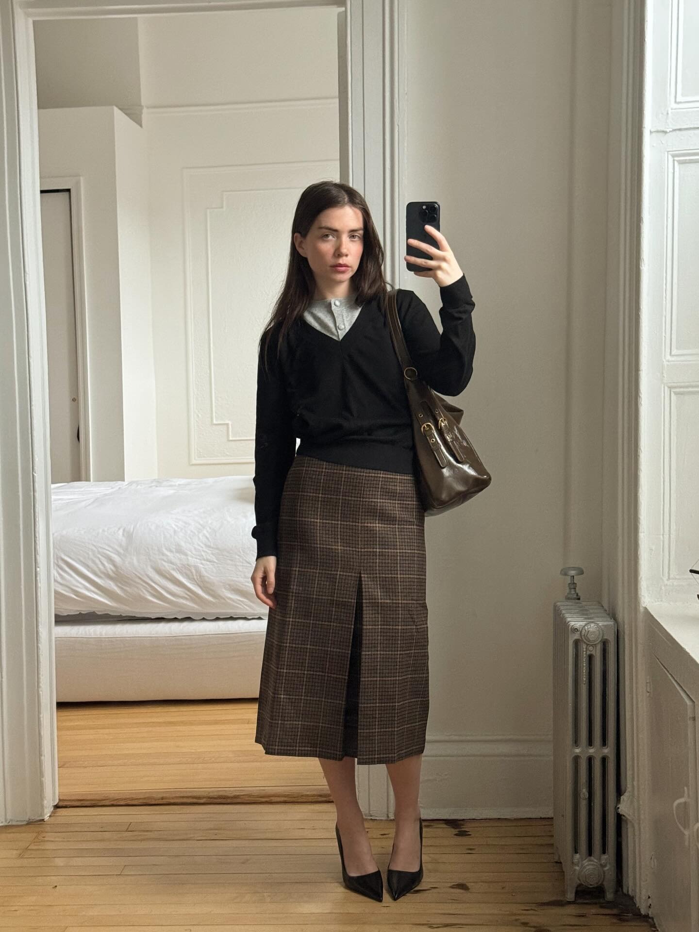 @accidentalinfluencer wearing a skirt outfit idea for 2026, including a V-neck sweater with a plaid midi skirt.