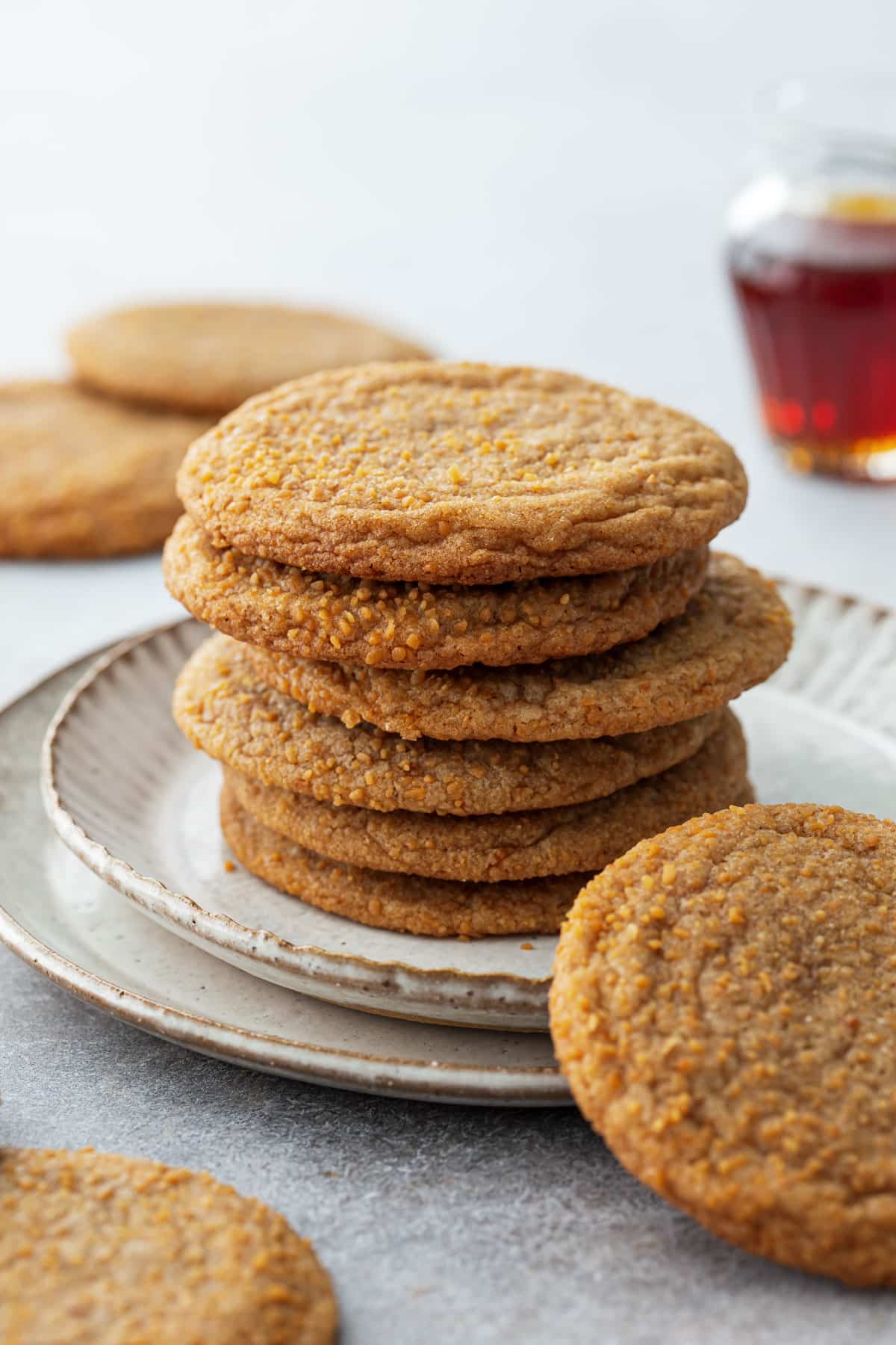 Stack of Chewy Maple Brown Butter Sugar Cookies on a ceramic plate, with a small glass pitcher of maple syrup and more cookies in the background.